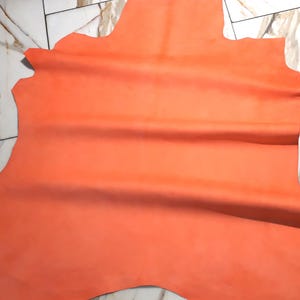 May include: A large, irregularly shaped piece of orange-colored leather. The leather has a smooth, matte finish and appears to be a single, uncut hide. The edges are uneven, suggesting it's been cut from a larger piece. The leather is likely used for crafting.