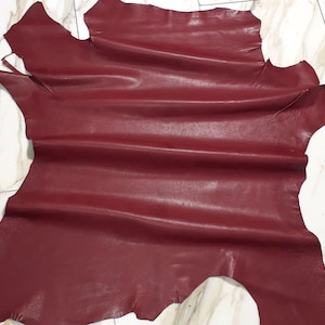 May include: A large, irregularly shaped piece of burgundy leather. The leather has a smooth, slightly textured surface with natural variations in colour and tone. The edges are uneven, indicating it's a cut piece. Suitable for various crafting projects.