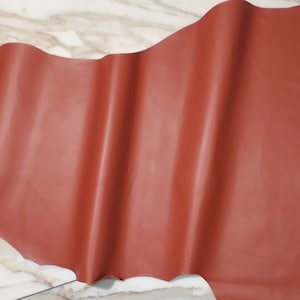 May include: A large, irregularly shaped piece of reddish-brown leather. The leather has a smooth, slightly glossy surface with subtle variations in color and texture. The material appears to be flexible and pliable, suitable for crafting.