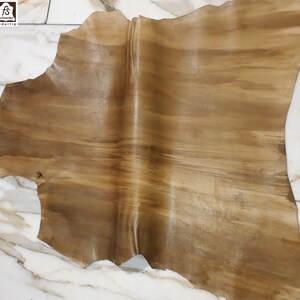 Puede incluir: Un trozo de cuero curtido con un patrón natural de vetas de madera en tonos marrones y beige. El cuero tiene un acabado liso y brillante y bordes irregulares. El logotipo de Schneider Ledertip está en la esquina superior izquierda.