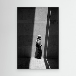 Puede incluir: Una fotografía en blanco y negro que muestra a una persona de pie contra un gran muro de piedra. El individuo está situado dentro de un haz vertical de luz brillante que contrasta con las sombras oscuras circundantes, vistiendo un pañuelo y pantalones.