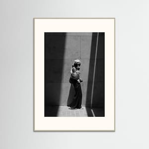 May include: A black and white framed wall art print featuring a person wearing a headscarf, a long-sleeved top, and long trousers, standing against a large stone-tiled wall. The scene is captured with high contrast, showing deep shadows and bright light.