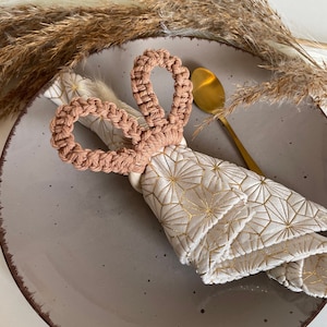 Könnte beinhalten: Ein hellrosa gehäkelter Hasen-Serviettenring auf einem grauen Teller mit einer weißen Serviette mit goldenem geometrischem Muster.