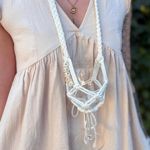 May include: A white macrame wine glass holder featuring a long braided strap and a net-like cradle for the glass. The holder is accented with light-colored wooden cube beads arranged vertically, each displaying a single black letter spelling BRAUT.
