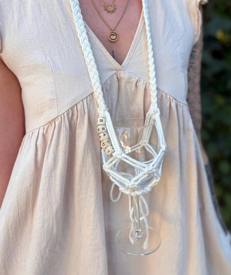 May include: A white macrame wine glass holder featuring a long braided strap and a net-like cradle for the glass. The holder is accented with light-colored wooden cube beads arranged vertically, each displaying a single black letter spelling BRAUT.