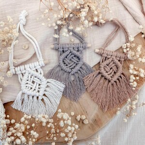 Könnte beinhalten: Drei Makramee-Wandbehänge in Weiß, Grau und Beige, jeweils mit geometrischem Design und Fransen. Die Aufhänger werden auf einer Holzoberfläche präsentiert und sind von kleinen weißen Blumen umgeben, was eine Boho-Ästhetik erzeugt.