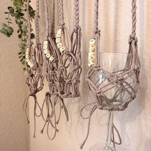 May include: A series of light gray macrame wine glass holders featuring braided straps and knotted netting. Each holder includes wooden beads with the printed names JACKY, LAURA, and SARAH, designed to suspend a clear glass stemware piece.