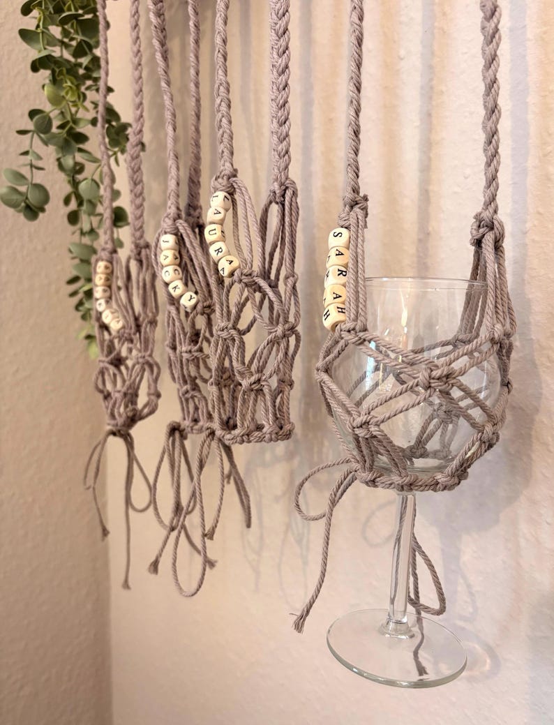 May include: A series of light gray macrame wine glass holders featuring braided straps and knotted netting. Each holder includes wooden beads with the printed names JACKY, LAURA, and SARAH, designed to suspend a clear glass stemware piece.