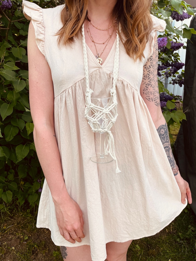 May include: A cream-colored macrame wine glass holder featuring a braided neck strap and a knotted net design that secures a clear glass goblet. The accessory is crafted from thick cotton cord and hangs at chest level, designed to hold a single glass.