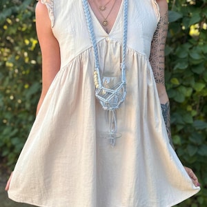 May include: A light beige sleeveless dress with ruffled cap sleeves and a V-neckline, paired with a light blue macrame wine glass holder necklace featuring a braided cord, a knotted net pouch, and small wooden beads hanging around the neck.