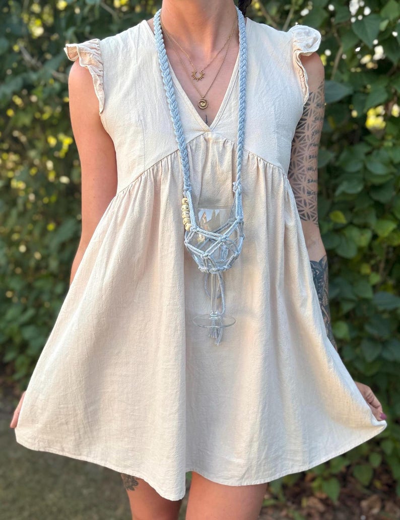 May include: A light beige sleeveless dress with ruffled cap sleeves and a V-neckline, paired with a light blue macrame wine glass holder necklace featuring a braided cord, a knotted net pouch, and small wooden beads hanging around the neck.