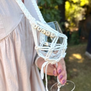May include: A white macrame wine glass holder featuring a braided strap and a net-like cradle for the glass. The holder is accented with wooden beads spelling out the word BRAUT in black capital letters, securely holding a clear glass stemware.