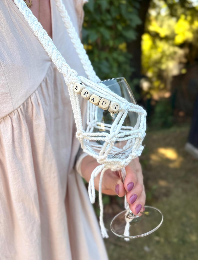 May include: A white macrame wine glass holder featuring a braided strap and a net-like cradle for the glass. The holder is accented with wooden beads spelling out the word BRAUT in black capital letters, securely holding a clear glass stemware.