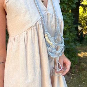 May include: A light blue macrame wine glass holder featuring a long braided strap and a net-like cradle. The holder is adorned with small square wooden beads spelling out the name MATTI in black capital letters, securely holding a clear glass.