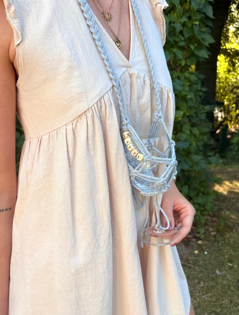 May include: A light blue macrame wine glass holder featuring a long braided strap and a net-like cradle. The holder is adorned with small square wooden beads spelling out the name MATTI in black capital letters, securely holding a clear glass.