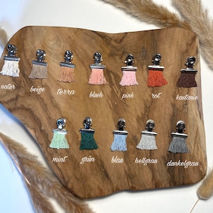 May include: A collection of twelve fabric tassels attached to silver metal clips, arranged in two rows on a wooden board. The tassels are in various colors including white, beige, terracotta, blush, pink, orange, brown, mint, green, blue, light gray, and dark gray.