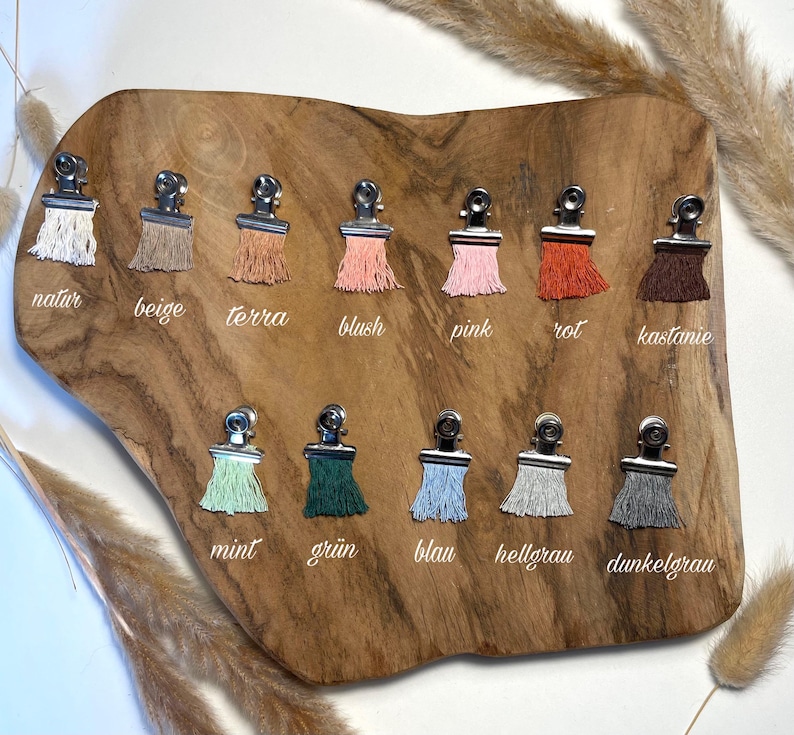 May include: A collection of twelve fabric tassels attached to silver metal clips, arranged in two rows on a wooden board. The tassels are in various colors including white, beige, terracotta, blush, pink, orange, brown, mint, green, blue, light gray, and dark gray.
