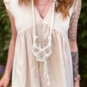 May include: A cream-colored macrame wine glass holder featuring a braided neck strap and a knotted net design that secures a clear glass. The holder is worn over a light beige dress with flutter sleeves and a V-neckline, paired with layered gold necklaces.