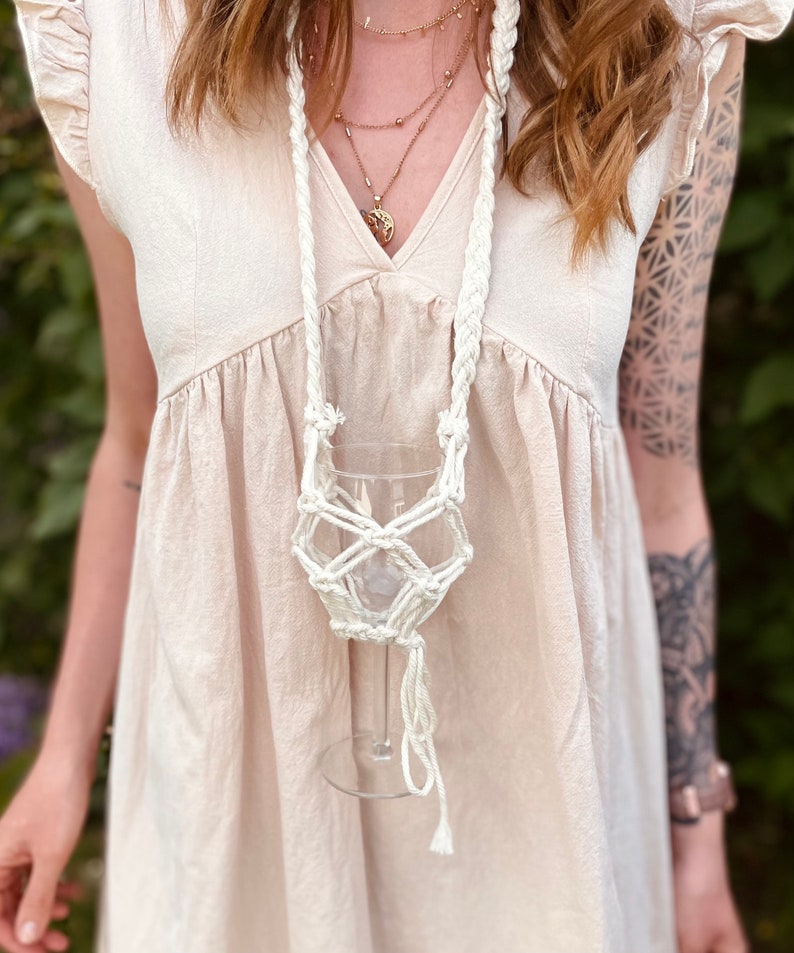 May include: A cream-colored macrame wine glass holder featuring a braided neck strap and a knotted net design that secures a clear glass. The holder is worn over a light beige dress with flutter sleeves and a V-neckline, paired with layered gold necklaces.