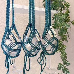 May include: Three clear glass wine glasses suspended in dark teal macrame holders. Each holder features braided cords and light-colored square beads with the black printed text CATYS JGA. The knotted cord design secures each glass base.
