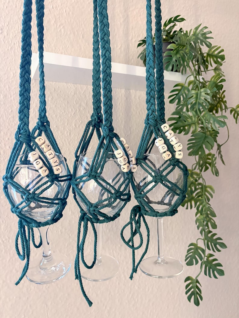 May include: Three clear glass wine glasses suspended in dark teal macrame holders. Each holder features braided cords and light-colored square beads with the black printed text CATYS JGA. The knotted cord design secures each glass base.