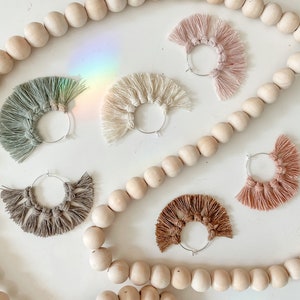 May include: Six pairs of handmade hoop earrings with fringe tassels in various colors, including beige, brown, green, and pink. The earrings are displayed on a white surface with wooden beads.