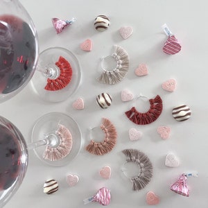 May include: A flat lay of Valentine's Day themed wine glass charms. The charms are made of fabric with fringe and come in three colors: pink, red, and beige. The charms are arranged around wine glasses, chocolate hearts, and candy hearts. The candy hearts have messages like "Treat Me", "I Love You", and "Hug Me".