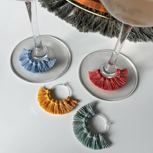 May include: Four wine glass charms with colorful tassels. The charms are made of yarn and are attached to silver rings. The tassels are blue, red, yellow, and green.