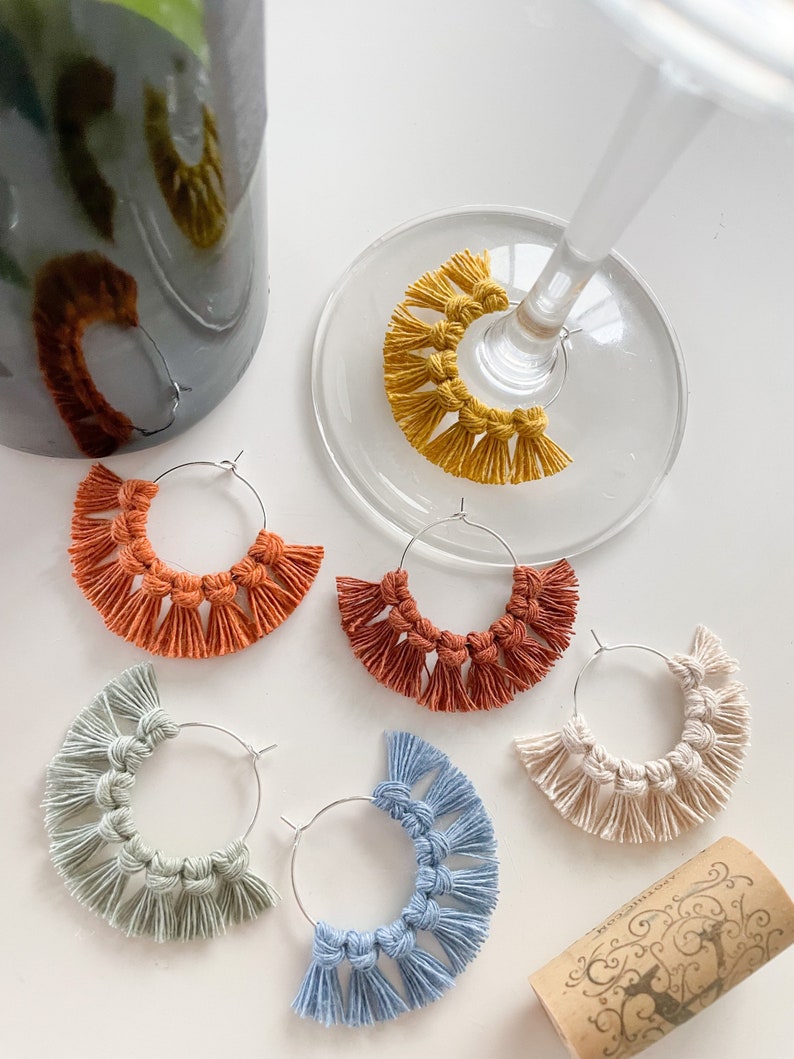 May include: Five pairs of handmade macrame earrings with fringe. The earrings are in various colors, including orange, red, blue, green, and yellow. The earrings are hanging from silver hoops.