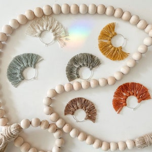 May include: Six pairs of handmade macrame earrings in various colors, including cream, green, yellow, brown, and orange. The earrings are displayed on a white surface with a string of wooden beads.