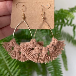 Puede incluir: Un par de pendientes de aro dorados con flecos marrones. Los pendientes están hechos de un fino alambre dorado y tienen forma de lágrima. Los flecos están hechos de un material suave y marrón y están anudados en la parte superior.