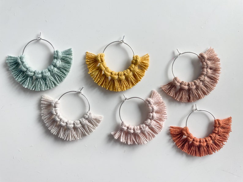 May include: Six pairs of handmade hoop earrings with fringe. The earrings are made with macrame and come in a variety of colors, including light blue, yellow, pink, and orange.