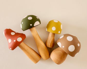 May include: Four wooden mushroom figurines with painted caps in red, green, yellow, and brown. Each mushroom cap has white polka dots. The stems are made of natural wood. The mushrooms are arranged on a light beige surface.