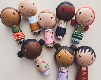 May include: A collection of eight hand-painted wooden peg dolls featuring diverse skin tones, hair colors, and styles. Each figure has a rounded head with a simple facial expression and wears a unique outfit, including dresses, shirts, and overalls.