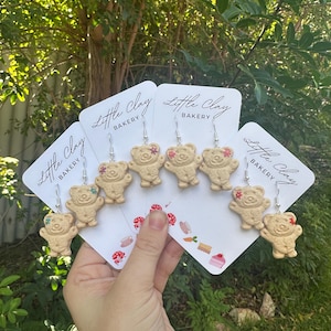 May include: Five pairs of clay bear earrings with pink and green details. The earrings are displayed on cards with the text "Little Clay Bakery".