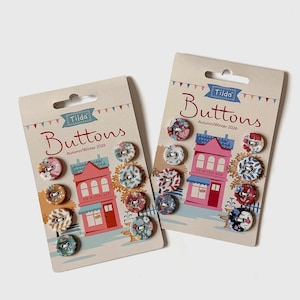 May include: Two rectangular cardboard display cards labeled Tilda Buttons Autumn/Winter 2026, each featuring a colorful illustration of a shop. Each card holds several small, round, fabric-covered buttons in various floral and geometric patterns.