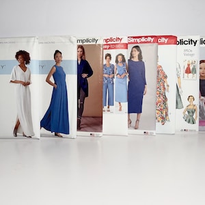 May include: A selection of Simplicity sewing pattern envelopes displaying images of dresses and other clothing items. The patterns feature diverse styles, including a white maxi dress, a blue dress, and vintage designs. The envelopes are arranged next to each other.