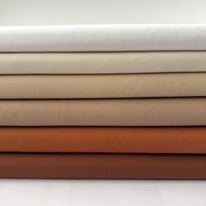 May include: A stack of fabric in various shades of neutral colors, including white, beige, tan, and brown. The fabric appears to be smooth and tightly woven, with a slight sheen. The colors range from light to dark, creating a gradient effect.