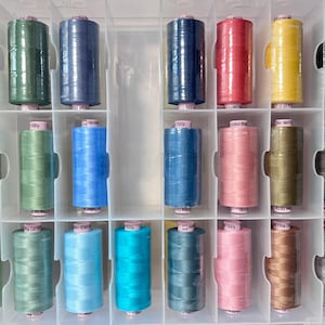 May include: A clear plastic storage box filled with spools of thread in various colors, including green, blue, yellow, red, and gray. The spools are labeled with different brand names and numbers.