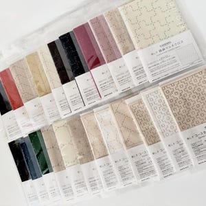 May include: Assortment of Cosmo brand cotton linen fabric squares in various colors, including red, black, and beige. Some squares feature stitched geometric patterns. The packaging includes Japanese text and measurements.