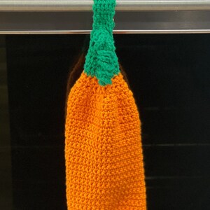 May include: A handmade, orange crochet kitchen towel shaped like a pumpkin, with a green stem and loop for hanging. The towel is hanging on a stainless steel oven handle. The towel is a seasonal autumn decoration.