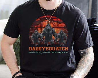 May include: A black t-shirt with a graphic of three Bigfoot figures in a forest setting. The text "Daddy'squatch" is printed below the figures, with the tagline "Like a Daddy, just way more Squatchy".