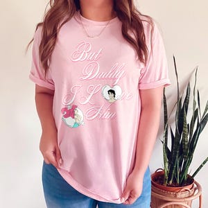 May include: Pink t-shirt with a graphic print of Ariel and Prince Eric from The Little Mermaid. The text on the shirt reads "But Daddy I Love Him."