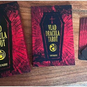 May include: A red and black tarot deck with a coffin design and the text "Vlad Dracula Tarot" on the front. The deck is laid out on a wooden surface.