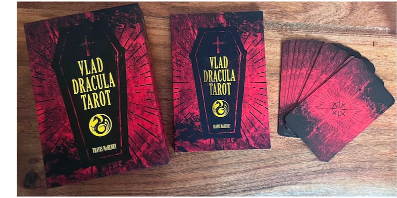 May include: A red and black tarot deck with a coffin design and the text "Vlad Dracula Tarot" on the front. The deck is laid out on a wooden surface.