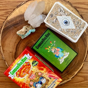 May include: Two boxes of Garbage Pail Kids Tarot Deck and Guidebook, one green and one red, with a white crystal cluster and a turquoise stone on a wooden surface.