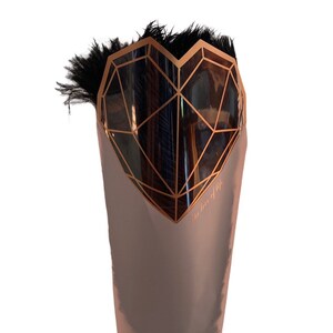 Puede incluir: Una bolsa de regalo de color oro rosa met&aacute;lico con un dise&ntilde;o de coraz&oacute;n geom&eacute;trico en cobre. El coraz&oacute;n est&aacute; lleno de plumas negras. Las palabras "the love of life" est&aacute;n impresas en el lateral de la bolsa.