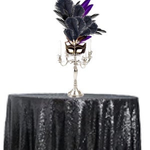 Puede incluir: Un candelabro plateado con cuatro velas encendidas, coronado con una m&aacute;scara de mascarada decorativa y plumas negras y moradas, se encuentra sobre un mantel negro con lentejuelas. La m&aacute;scara es marr&oacute;n y dorada.