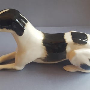 Puede incluir: Figura de perro de cerámica en posición de descanso. El perro tiene un acabado brillante con una combinación de colores blanco y negro. La cabeza, las orejas y la espalda son negras, el resto del cuerpo es blanco.