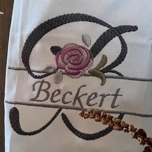 Könnte beinhalten: Weißes besticktes Handtuch mit floralem Design und dem Namen "Beckert". Die Stickerei zeigt eine Rose in Rosa- und Grüntönen mit einem großen Buchstaben "B" in Dunkelgrau. Das Handtuch wird vor einem rustikalen Holzhintergrund präsentiert.